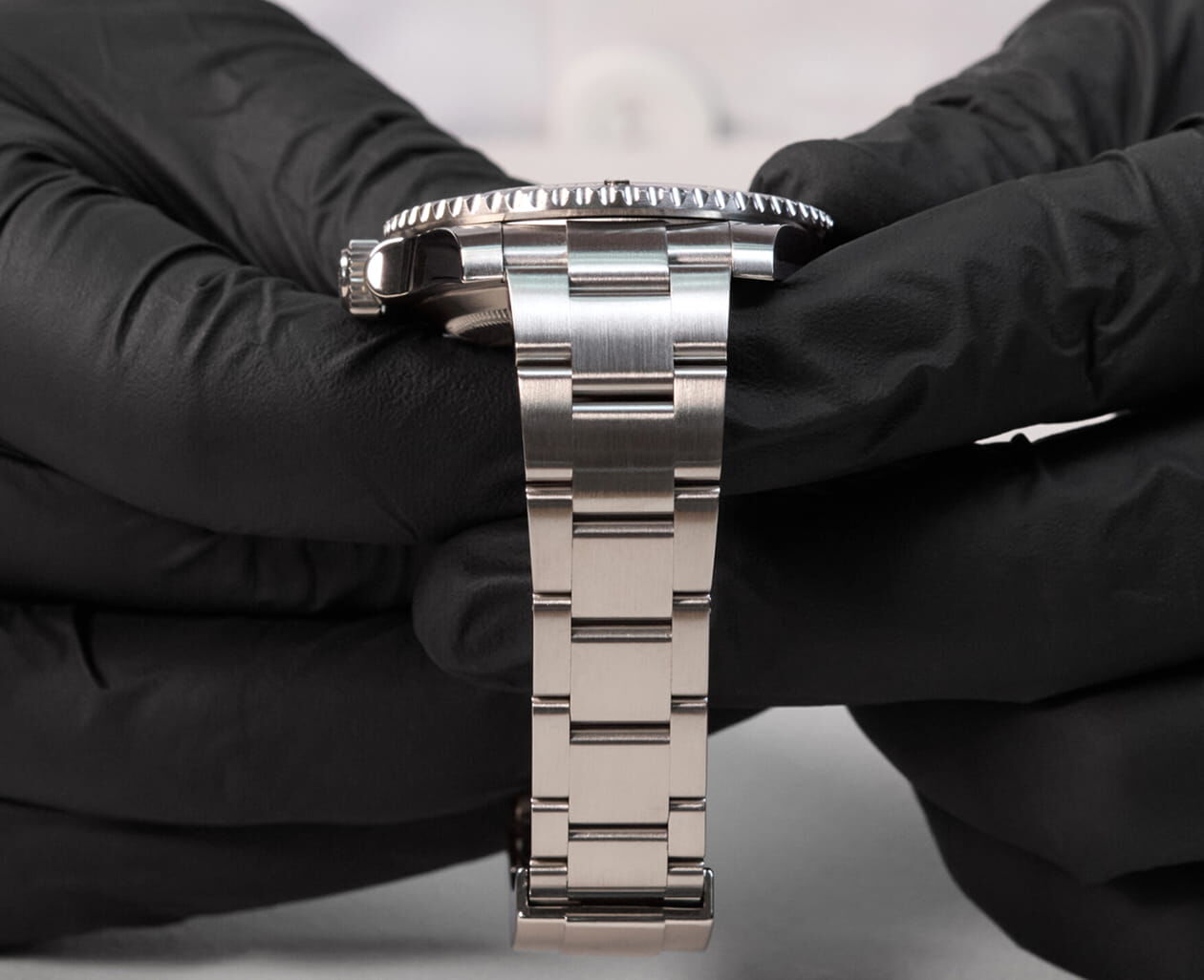 Someone wearing black gloves and a white lab coat inspects a stainless steel watch.