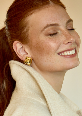 A woman smiles with eye slightly closed. She is wearing a pair of bold yellow gold earrings.