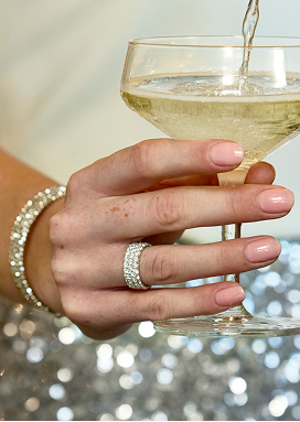 A woman&rsquo;s hand is shown holding a glass of champagne. She is wearing a diamond anniversary band and a sparkly bracelet.