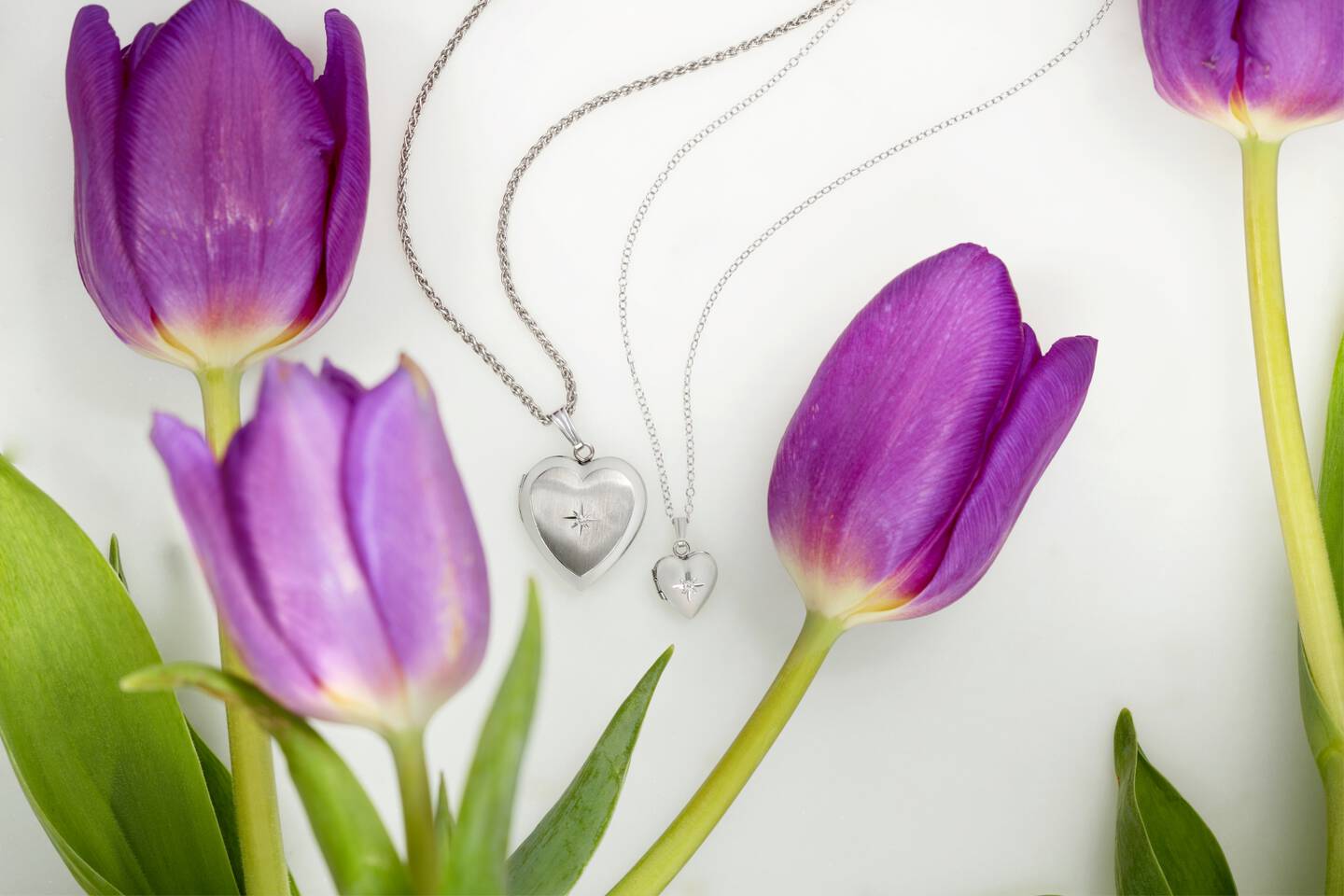 Matching mother and child silver heart pendants with a single diamond in the center shown amid purple tulips. 