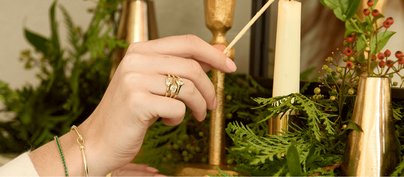 A hand wearing yellow gold and diamond rings lights a holiday candle amid a festive background of leaves and gilded candlesticks. 