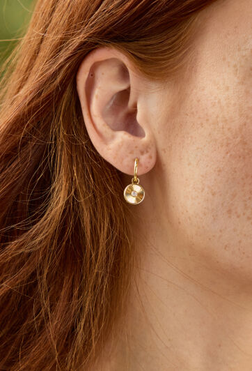 A woman in gold earrings and a cream colored coat turns to look at a camera.