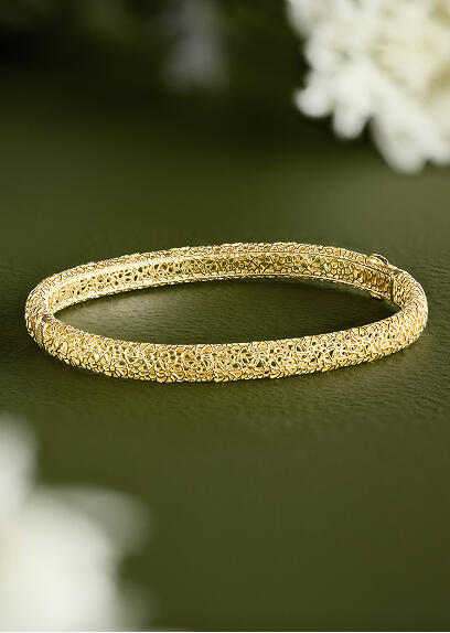 A yellow gold bangle rests atop a green surface with out-of-focus flower petals in the corner.