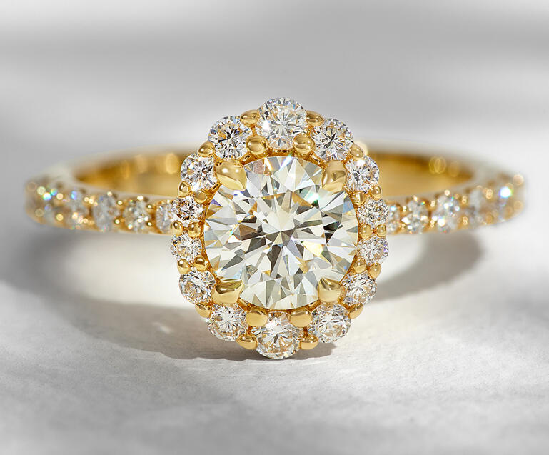 A diamond engagement ring in yellow gold featuring a halo of round diamonds and diamonds set along the shank. 