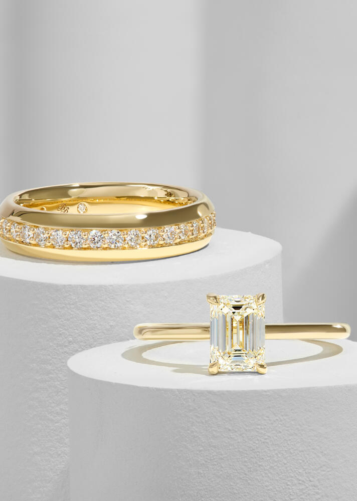 A solitaire engagement ring in yellow gold and a diamond band rest upon round white pillars.