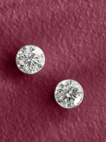 Round diamond stud earrings rest on a textured purple surface.