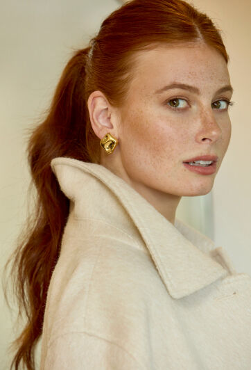 A woman in gold earrings and a cream colored coat turns to look at a camera.