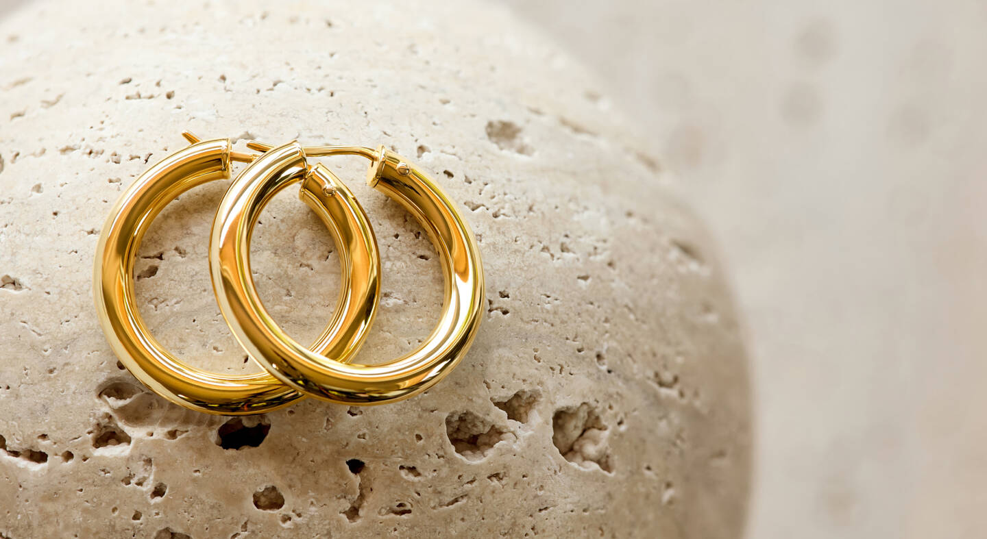 Two yellow gold hoop earrings overlap on a porous stone surface.