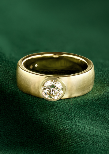 A wide wedding band with a solitaire diamond resting on its side on green fabric. 