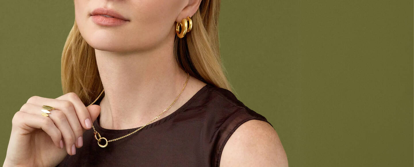 A woman wearing a tank top is touching her yellow gold necklace and wearing gold hoop earrings.
