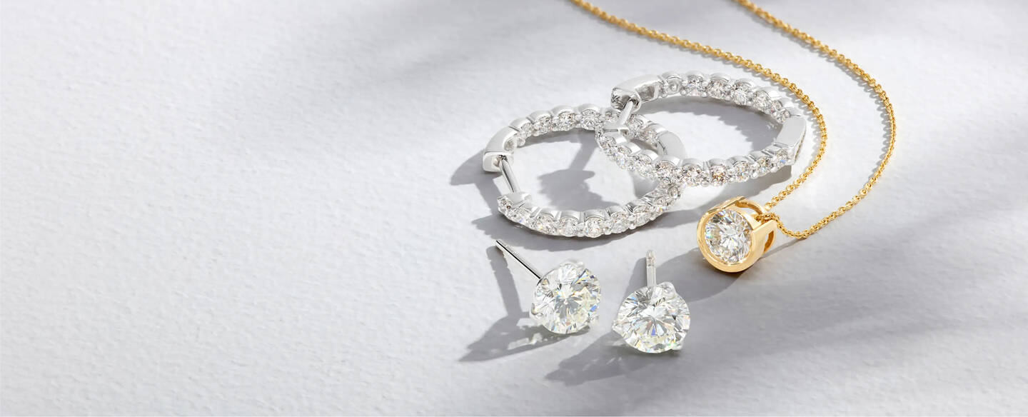Round diamond studs, hoops and a pendant in white and yellow gold settings sparkle against a light gray textured background.