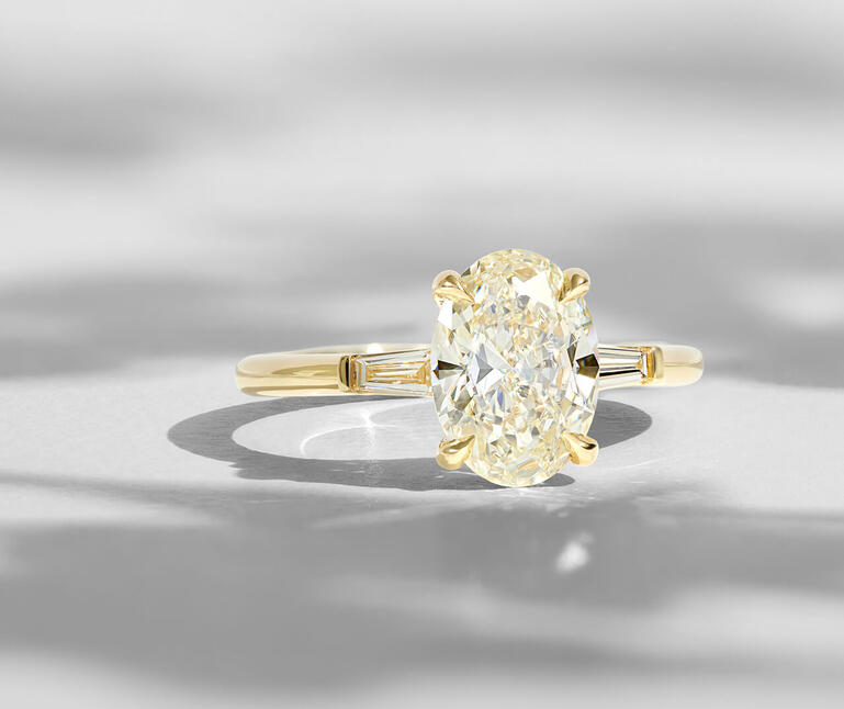 An oval diamond engagement ring in a prong setting of yellow gold with baguette accent diamonds along the shank. 