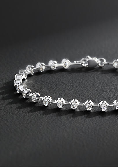  A ruby and diamond ring in 14k yellow gold on a charcoal colored background.  A yellow gold necklace smooth links on a smooth dark gray surface.  Round diamond stud earrings with a half-moon bezel setting of yellow gold.  A diamond bracelet in white gold sparkles against a dark gray surface.