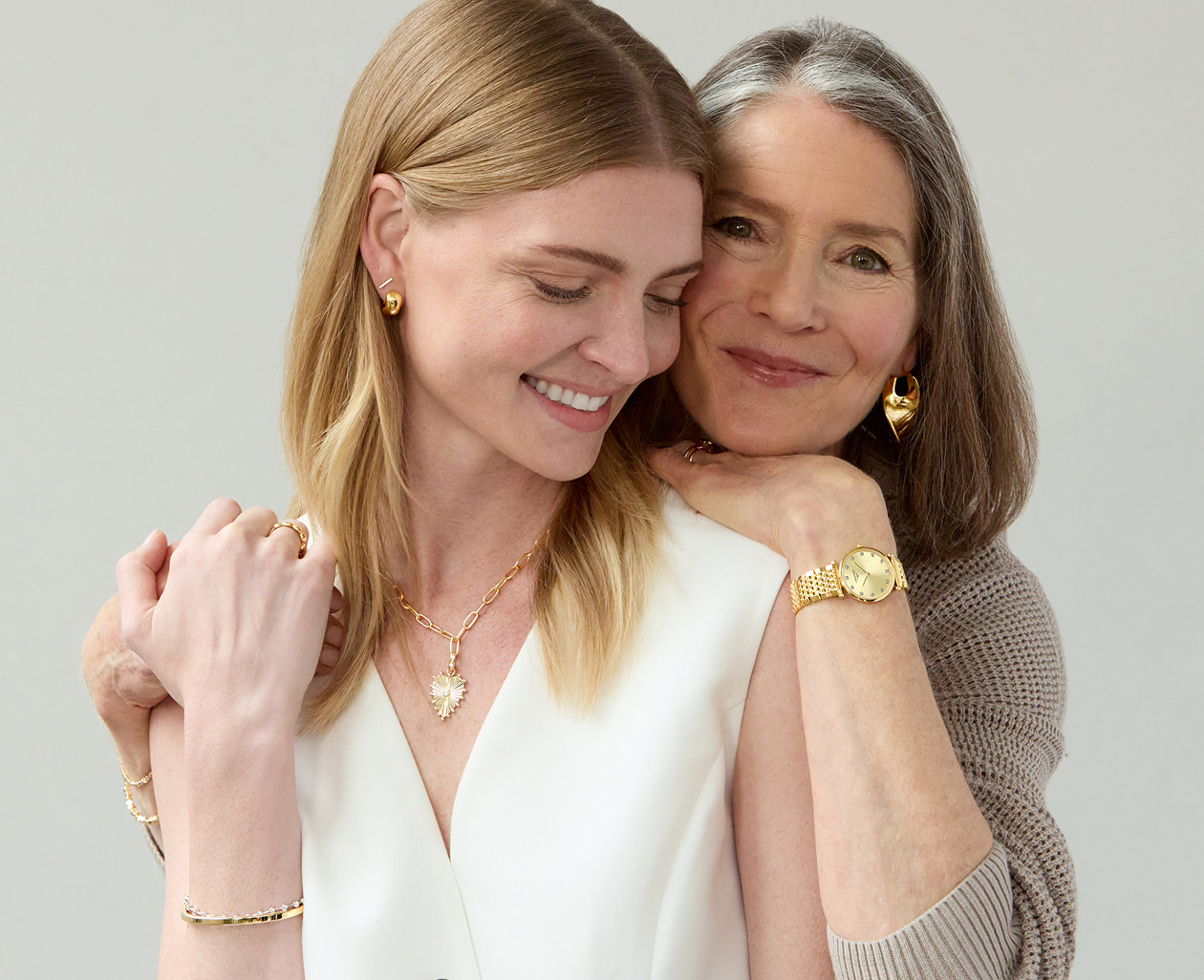 A mother embraces her adult daughter, both smiling and wearing yellow gold jewelry.