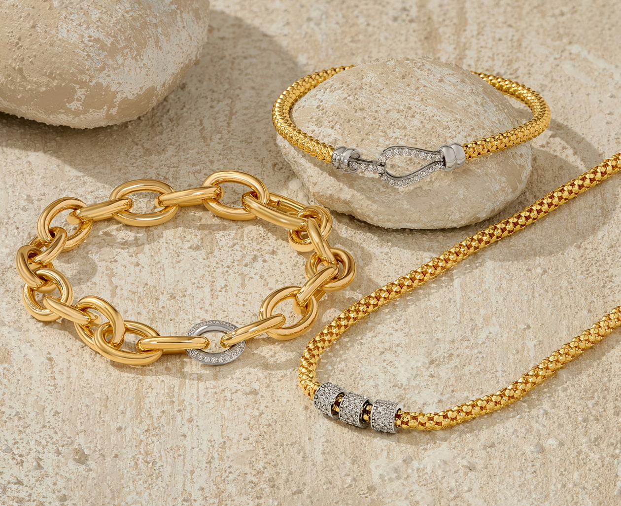 Two yellow gold bracelets and a yellow gold necklace with diamond accents are scattered across a light-colored stone surface.