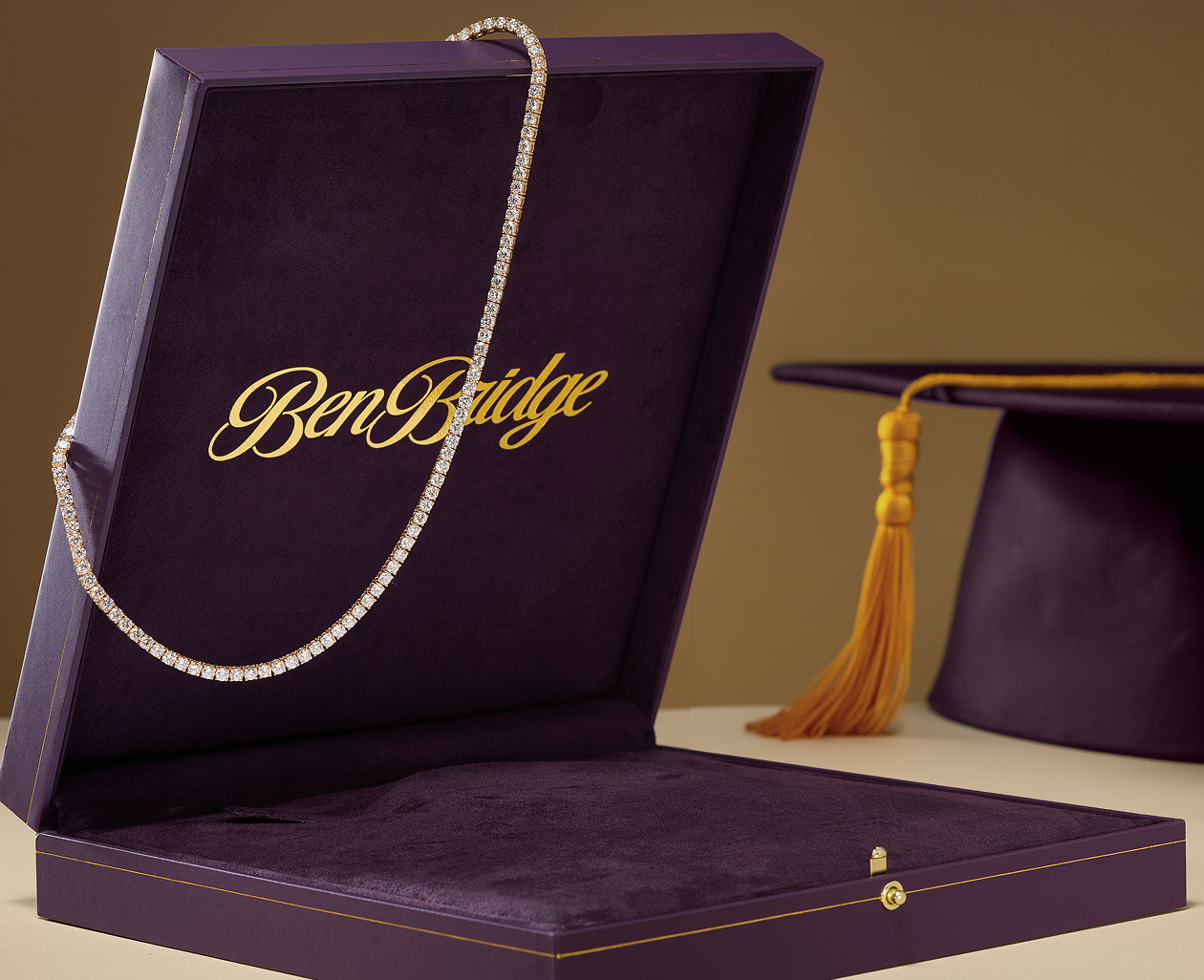 A diamond necklace hangs from the corner of a purple jewelry box alongside a purple graduation cap with a gold tassel.